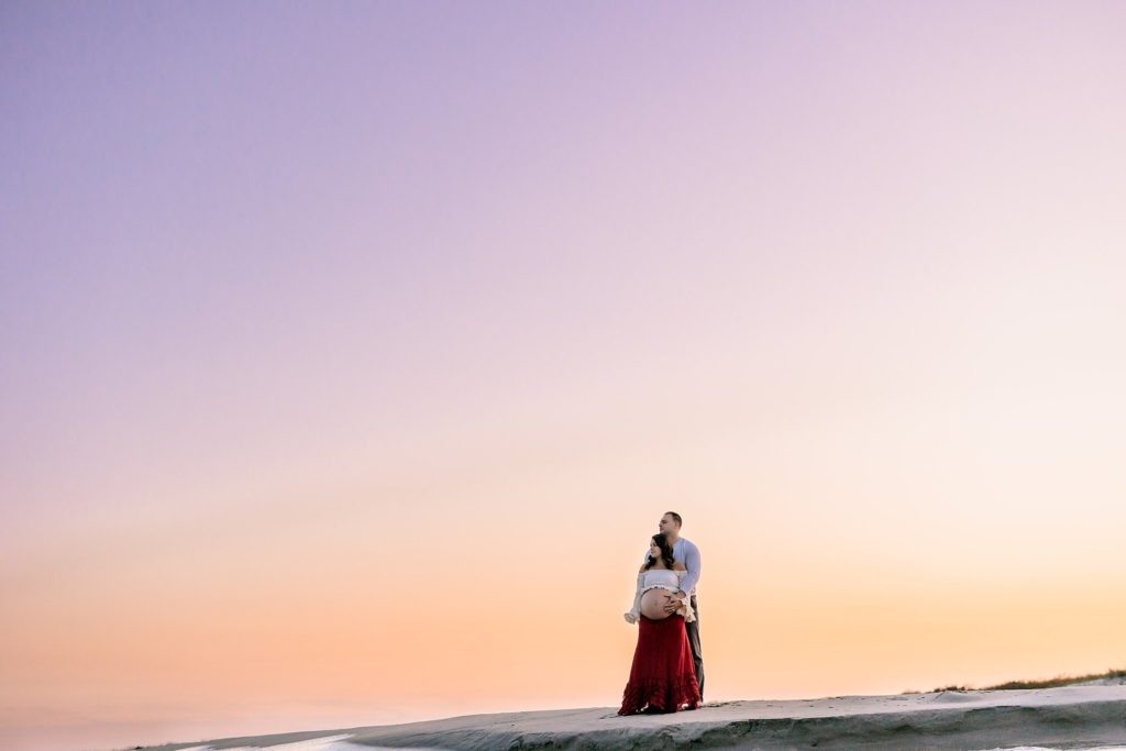 Pregnant 26 year old woman on the beach in Ocean City, New Jersey with sunset behind her in a ivory crop top and red skirt with her 29 year old husband